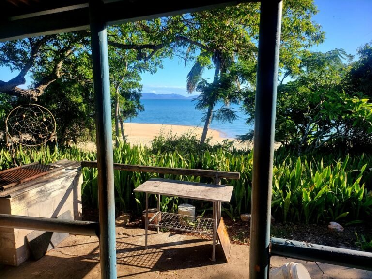 Sunset beach view from shaded bungalow patio with lush greenery and clear blue ocean.